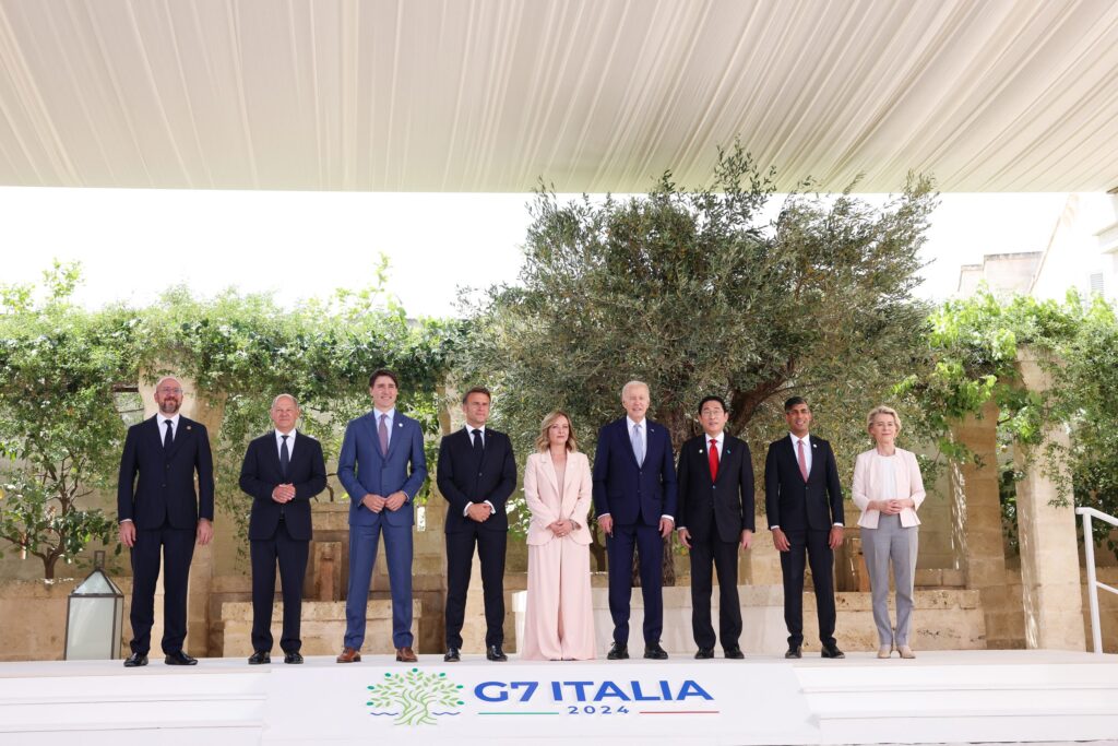 World leaders seated at a conference table during the 50th G7 summit in Fasano, Italy, with flags of member nations in the background.