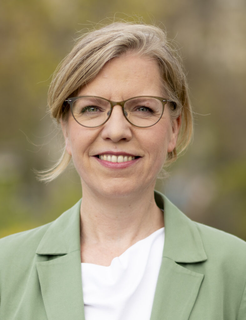 Leonore Gewessler speaking at a podium during the Green Party convention in Vienna, with delegates applauding in the background.