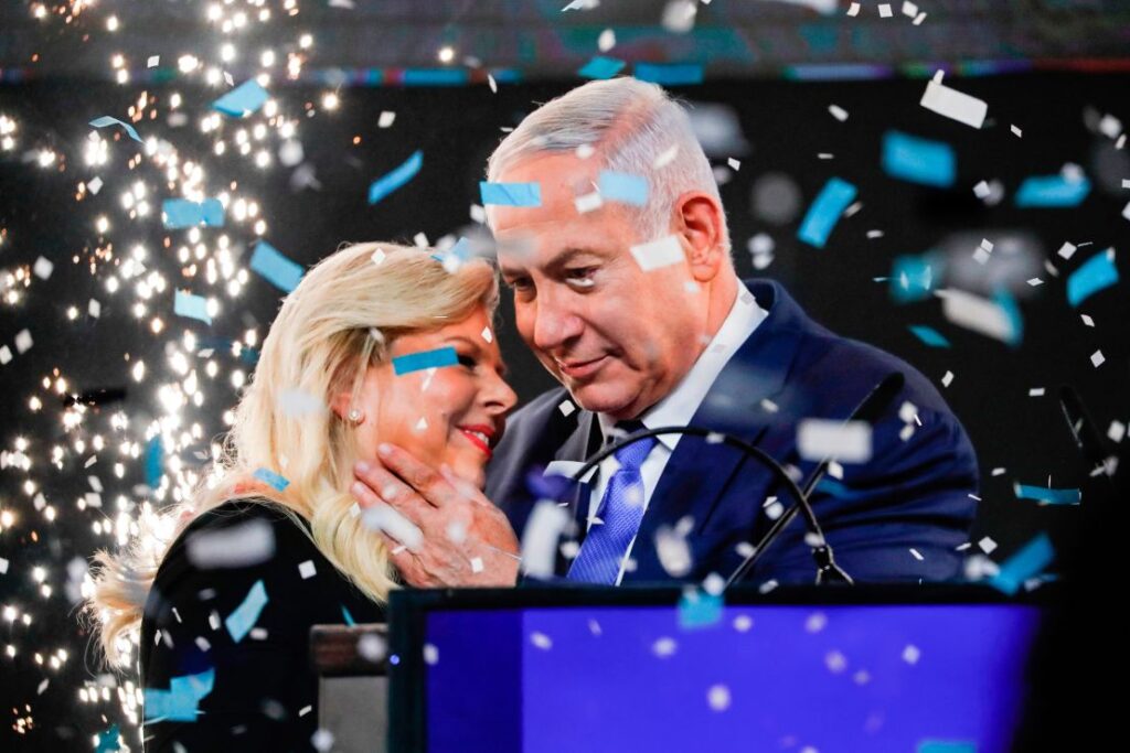 Benjamin Netanyahu speaks at a campaign event, surrounded by Likud party supporters holding blue and white flags.