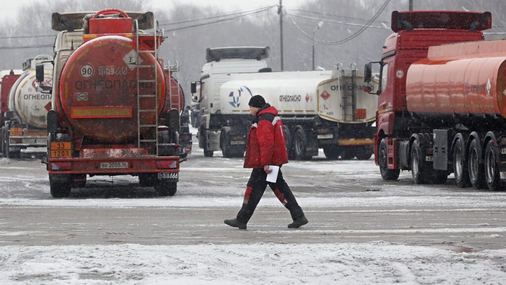 Rows of diesel pumps at an EU truck stop display prices while a cargo tanker flies the Russian flag in the background.