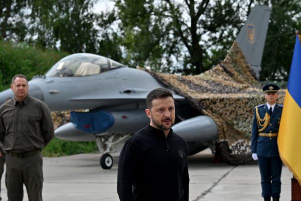 Ukrainian and Danish officials stand beside an F-16 fighter jet at a sunlit airbase press event.